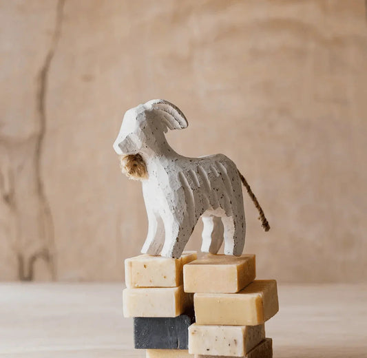 Hand-carved white wooden goat figurine with jute beard and tail, rustic farm decor, stacked soap blocks, heritage rural accent for American Farm Company lifestyle.