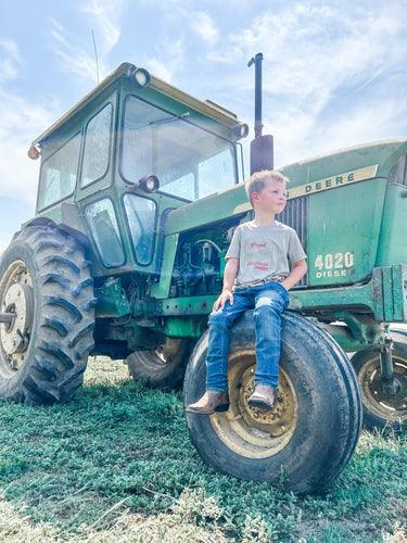 Young boy in distressed jeans and a heritage-style graphic tee sits on a vintage green tractor tire, embodying rural legacy and agricultural lifestyle for American Farm Company.