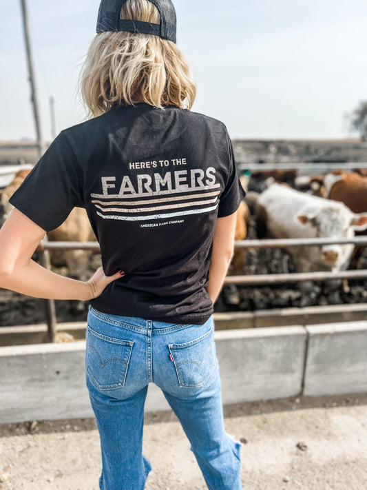 Rugged black graphic tee with vintage 'Here's to the Farmers' typography, relaxed fit, styled for rural women, paired with classic blue jeans at a cattle farm, embodying American Farm Company's agricultural heritage.
