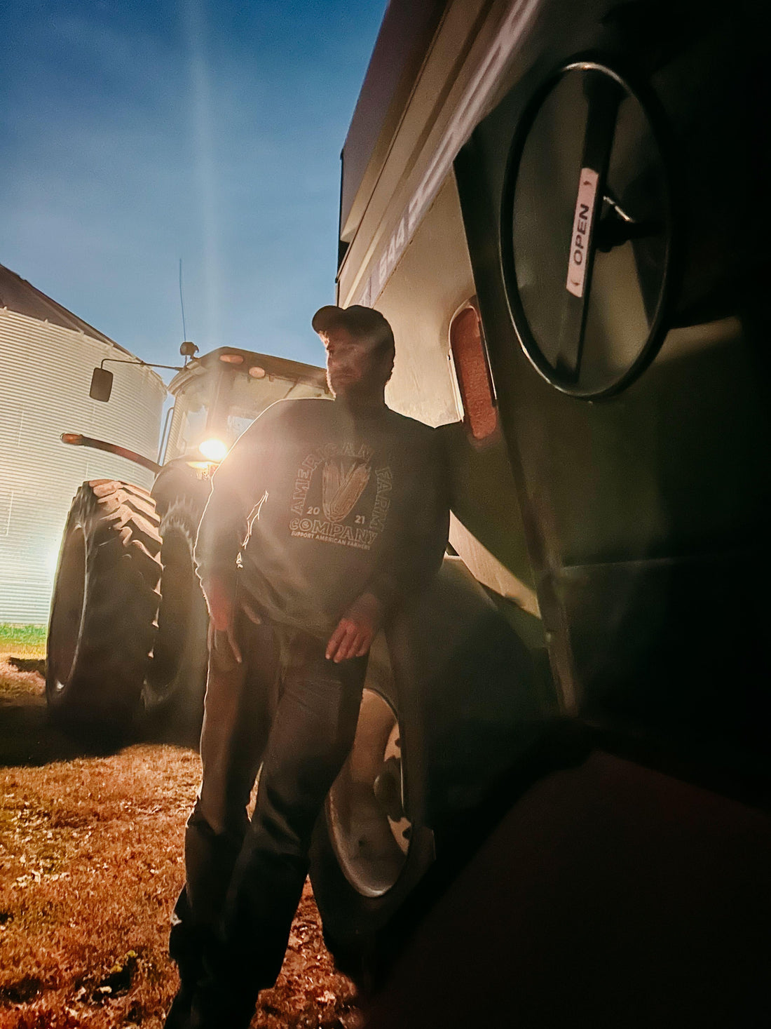 Rugged fit sweatshirt with heritage corn graphic and American Farm Company logo, worn by a man leaning against agricultural equipment at dusk, embodying rural legacy workwear style.
