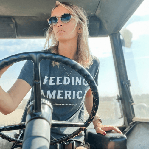 Charcoal relaxed-fit cotton tee with bold 'Feeding America' print, worn by woman driving tractor, rural heritage apparel by American Farm Company