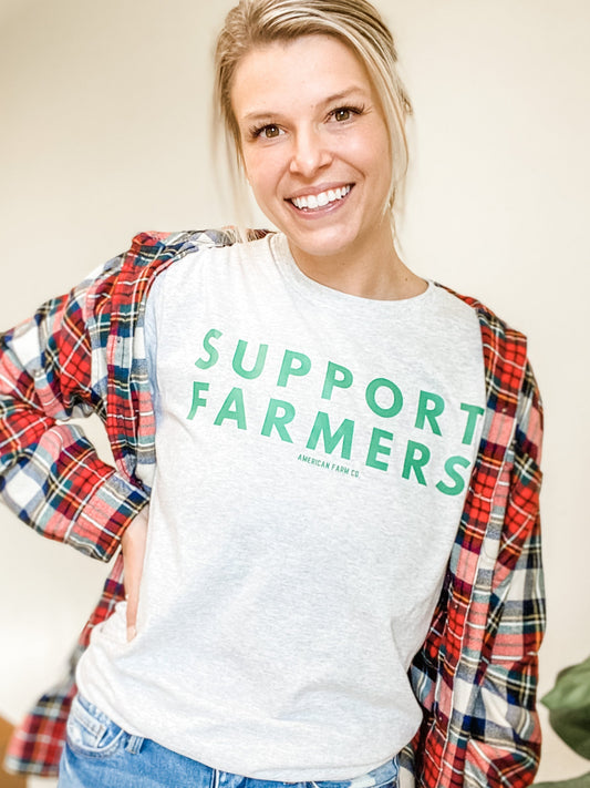 Heather gray relaxed fit tee with bold green SUPPORT FARMERS graphic, layered under a red plaid flannel, reflecting American Farm Company's heritage-inspired rural lifestyle apparel.