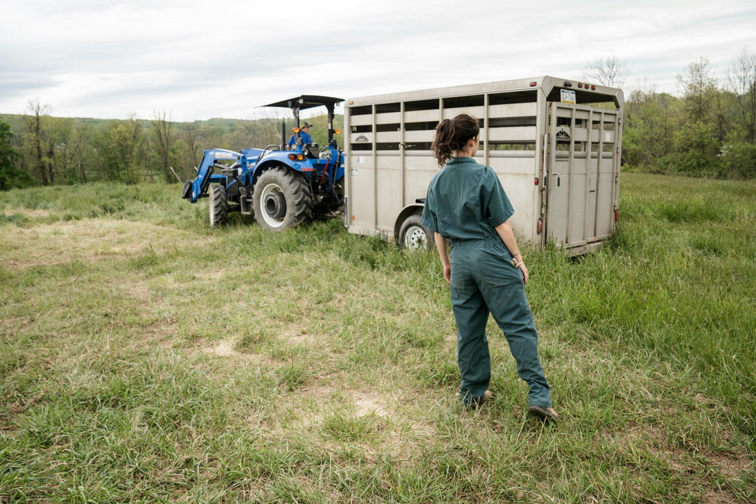 Rugged teal coveralls for women, relaxed fit agricultural workwear, shown beside livestock trailer and blue tractor in a heritage-inspired rural landscape