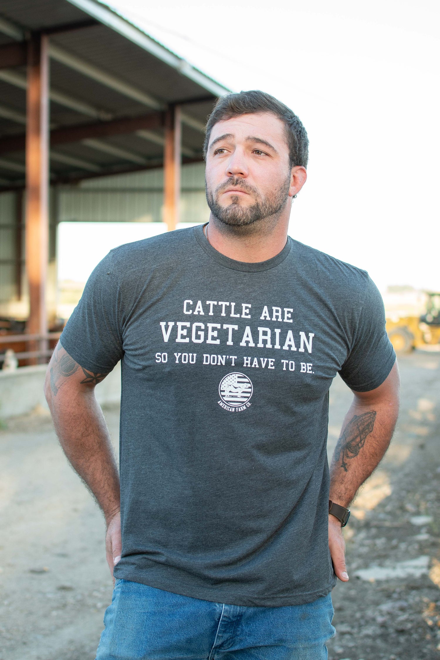 Heritage-inspired dark heather crewneck tee with bold 'Cattle Are Vegetarian' graphic, rugged rural workwear from American Farm Company, modeled outdoors on a farm.