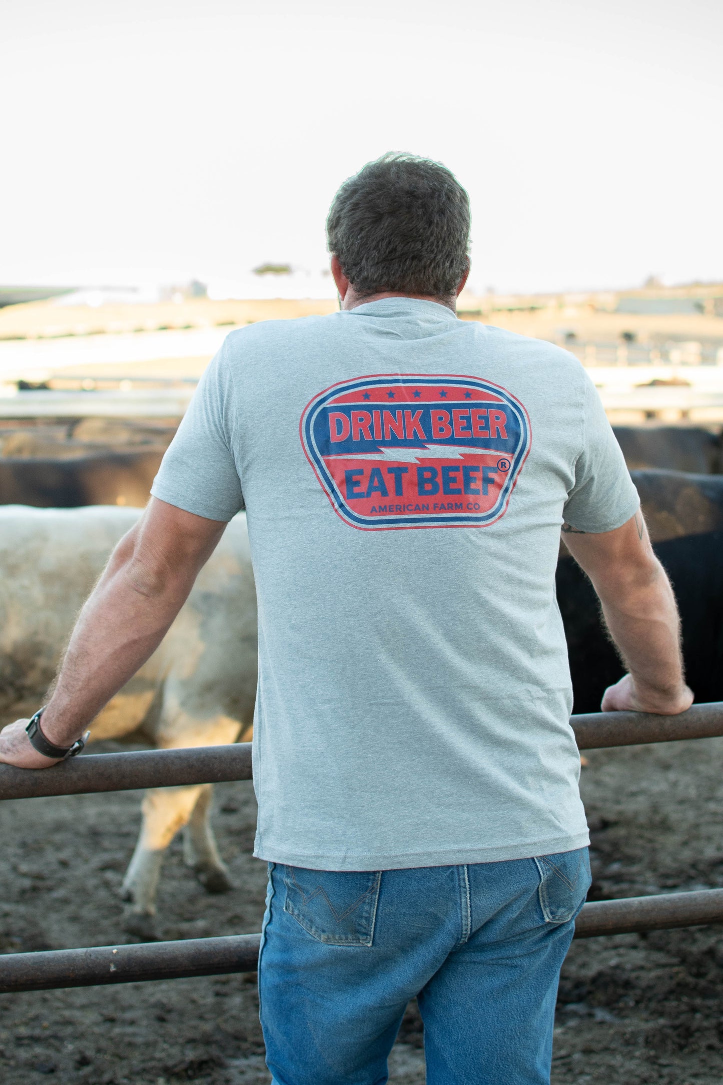 Heritage-inspired heather gray tee with bold 'Drink Beer Eat Beef' graphic, rural cattle farm setting, rugged American Farm Company workwear.