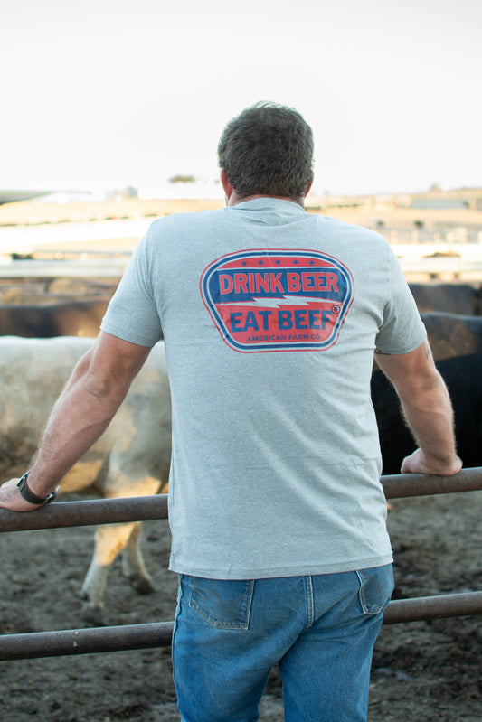 Heritage-inspired heather gray tee with bold 'Drink Beer Eat Beef' graphic, rural cattle farm setting, rugged American Farm Company workwear.
