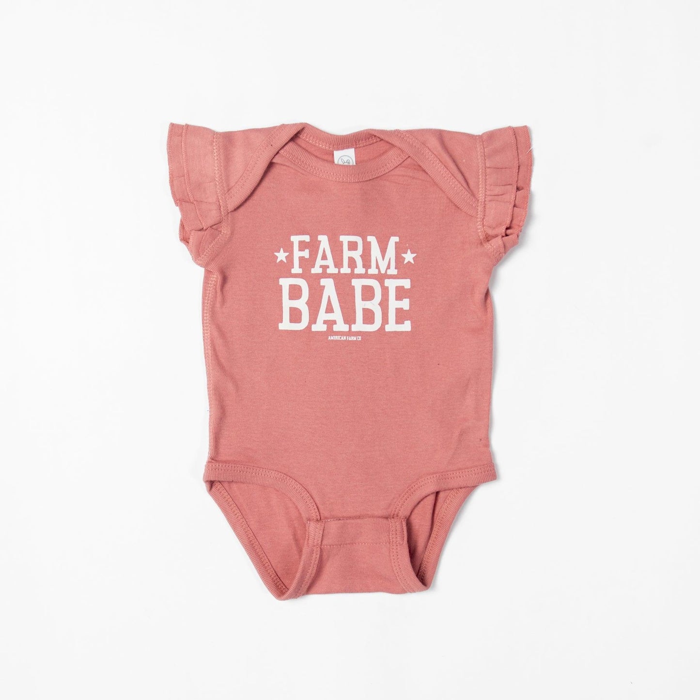 Heritage-inspired dusty rose baby onesie with ruffle sleeves, bold 'Farm Babe' graphic, and soft cotton fabric, embodying rural legacy from American Farm Company.
