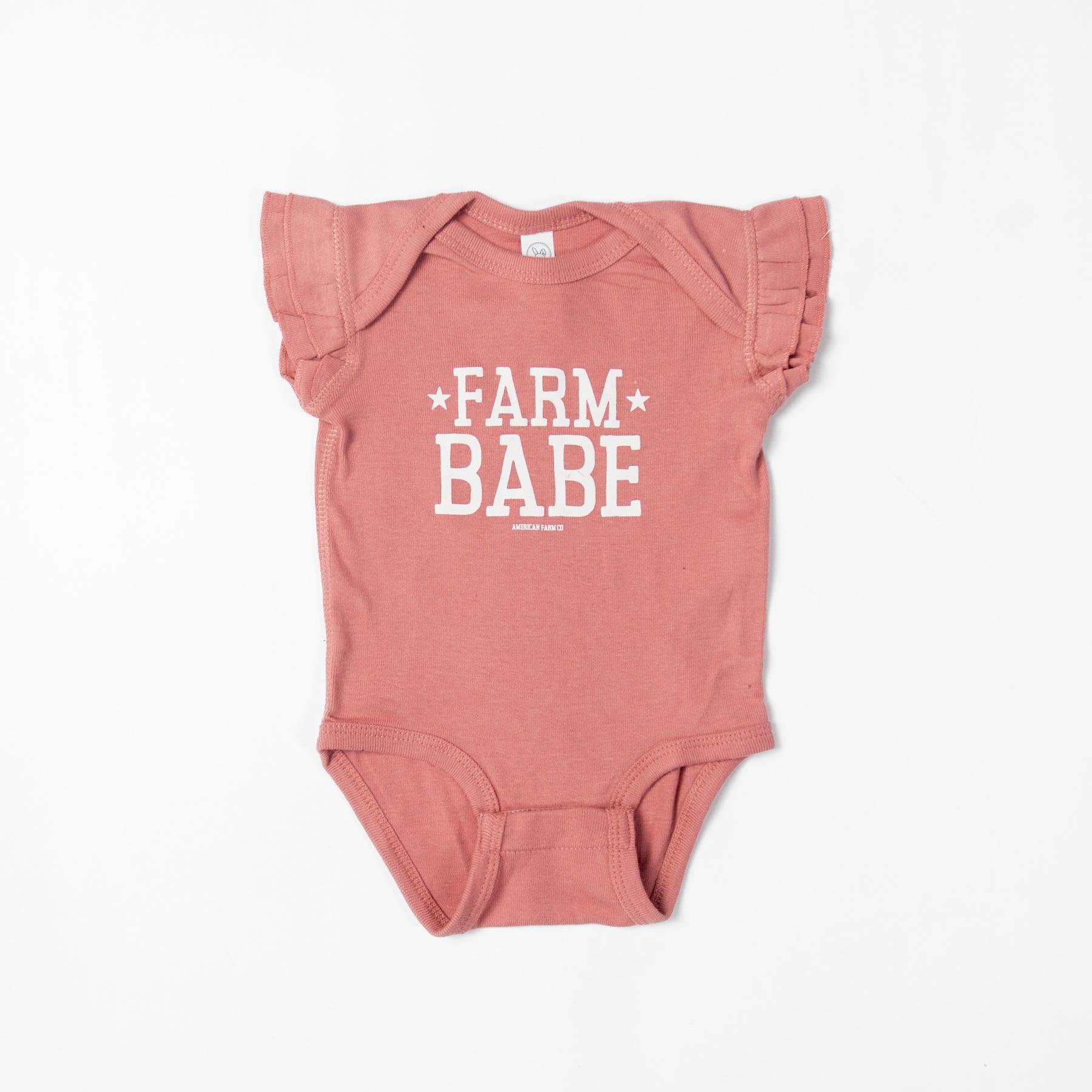 Heritage-inspired dusty rose baby onesie with ruffle sleeves, bold 'Farm Babe' graphic, and soft cotton fabric, embodying rural legacy from American Farm Company.