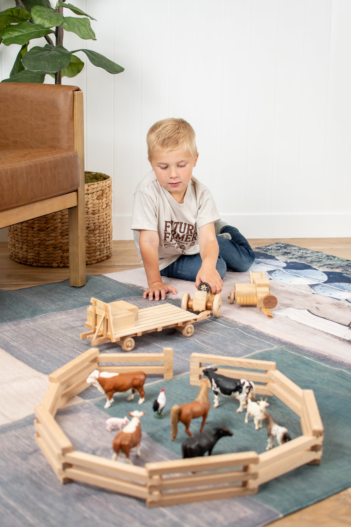 Blonde boy in relaxed-fit 'Future Farmer' tee and jeans playing with wooden farm toys and animal figurines on a rural farm-themed play rug, capturing American Farm Company’s heritage and agricultural legacy vibe.