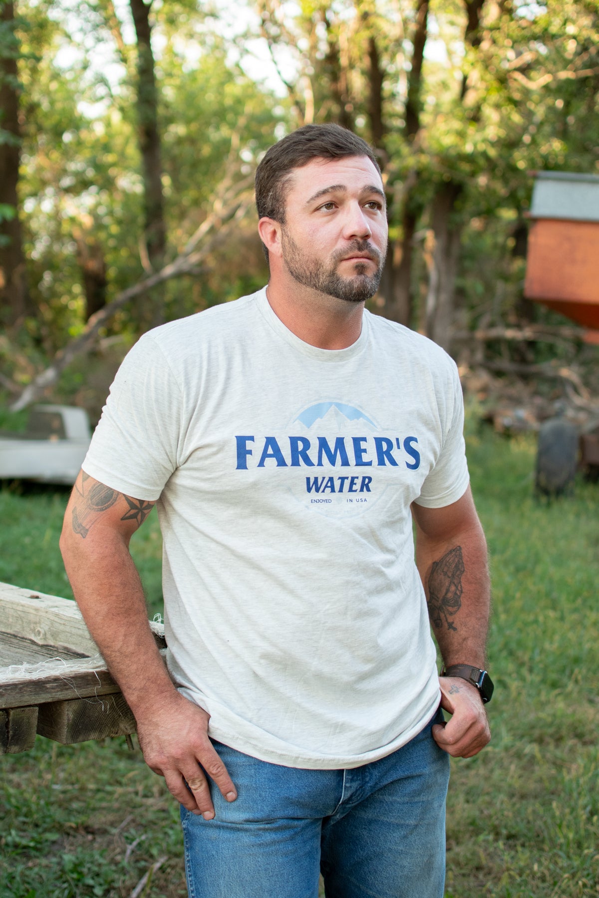 Heritage-inspired relaxed fit white tee with blue 'Farmer's Water' mountain graphic, classic American Farm Company rural workwear outdoors.
