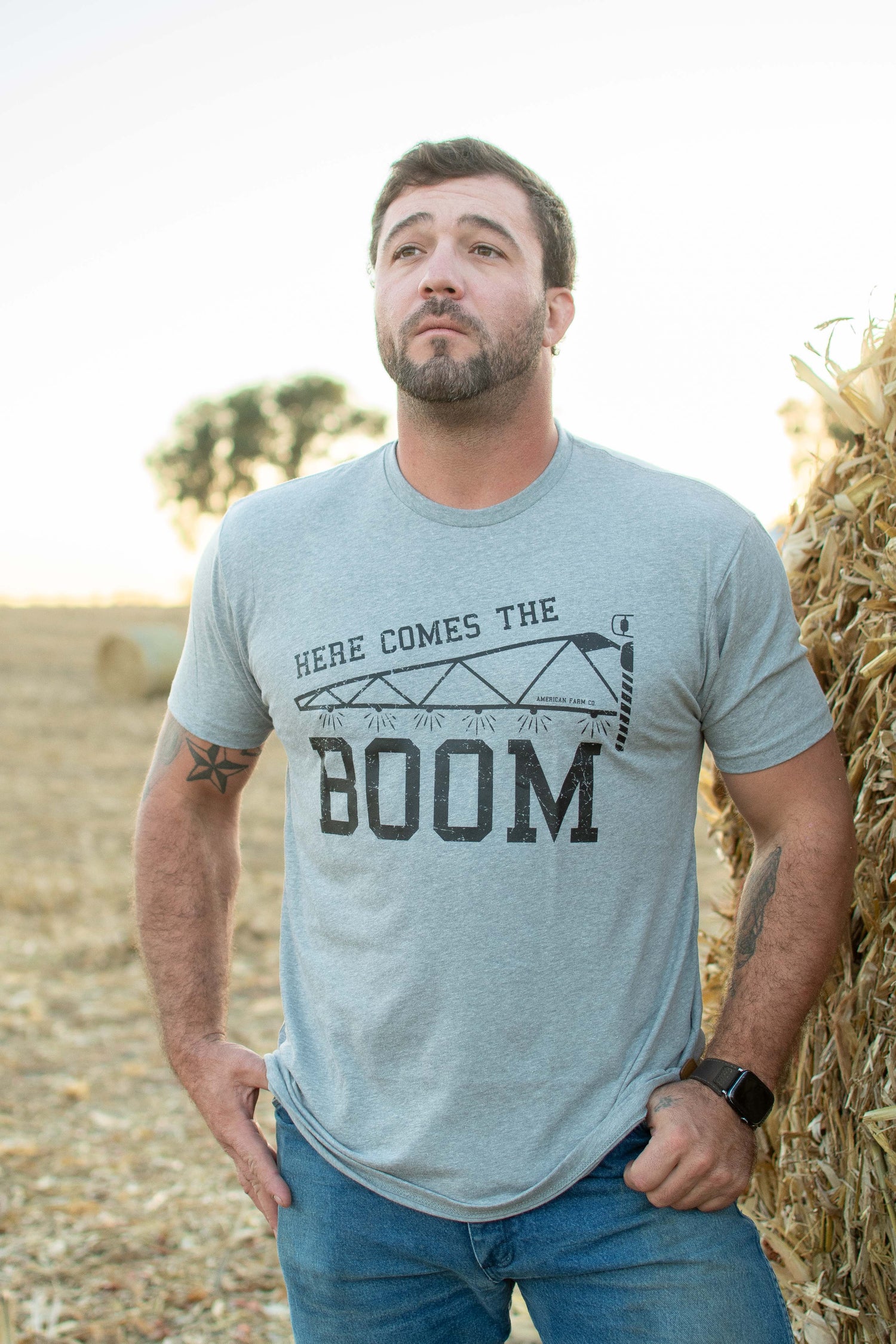 Heather gray relaxed fit tee with 'Here Comes the Boom' pivot irrigation graphic, rustic field backdrop, heritage rural workwear by American Farm Company.