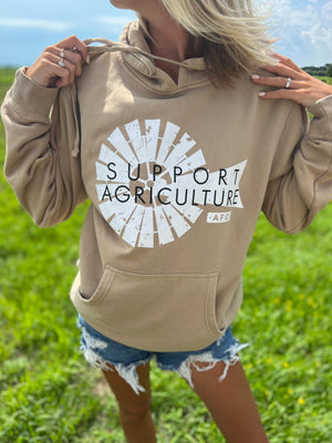 A woman in a field sporting a Support Agriculture Tan Hoodie paired with ripped shorts
