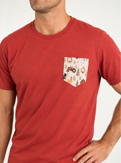 A man wearing a red crew-neck t-shirt with a decorative chest pocket featuring a whimsical autumn pattern of a red tractor, trees, and animals on a cream background.