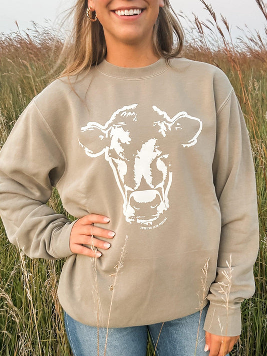 Heritage-inspired taupe crewneck sweatshirt with bold white cow graphic and 'American Farm Company' text, celebrating rugged rural lifestyle apparel.